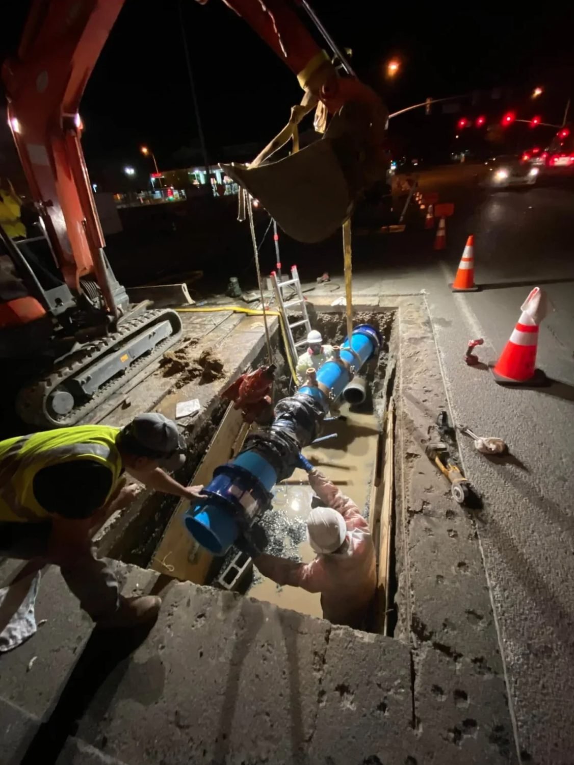 Night crew lowering a new water main section into a road excavation